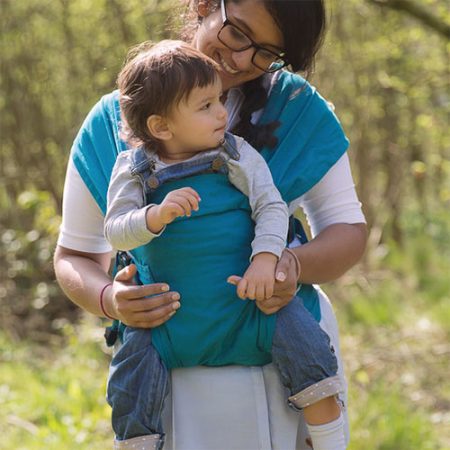 Woman carries older baby facing forwards in teal coloured Izmi Baby Carrier