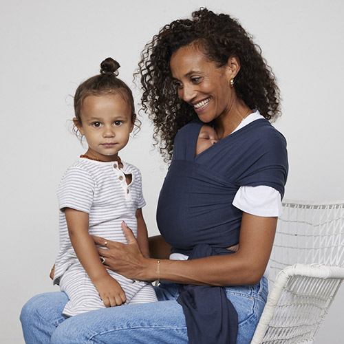 Seated woman carries baby on her front in Izmi Baby Wrap in Slate while toddler sits on her knee