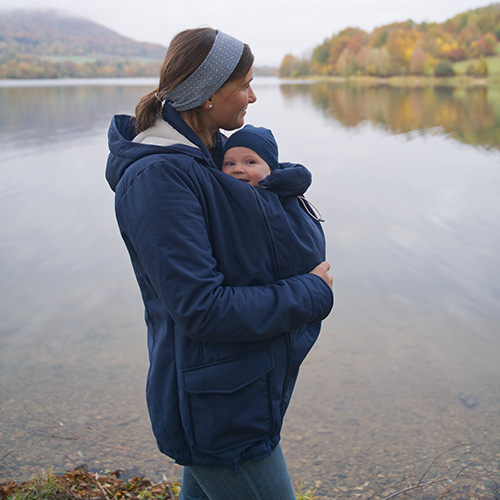 Woman carries baby on her front while wearing the Mamalila Cosy Allrounder babywearing coat in Navy