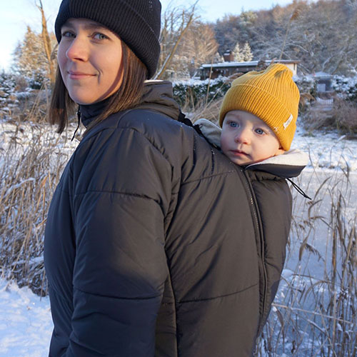 Woman carries baby on her back in baby carrier while wearing the Mamalila Copenhagen coat in black over the top of both of them