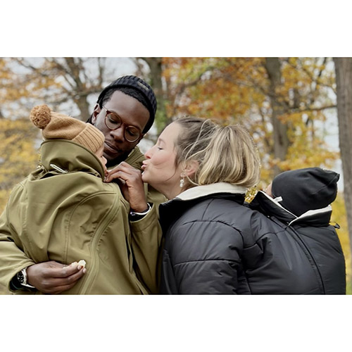 Man carries baby on his front while woman carries baby on her back; both wearing Mamalila babywearing coats